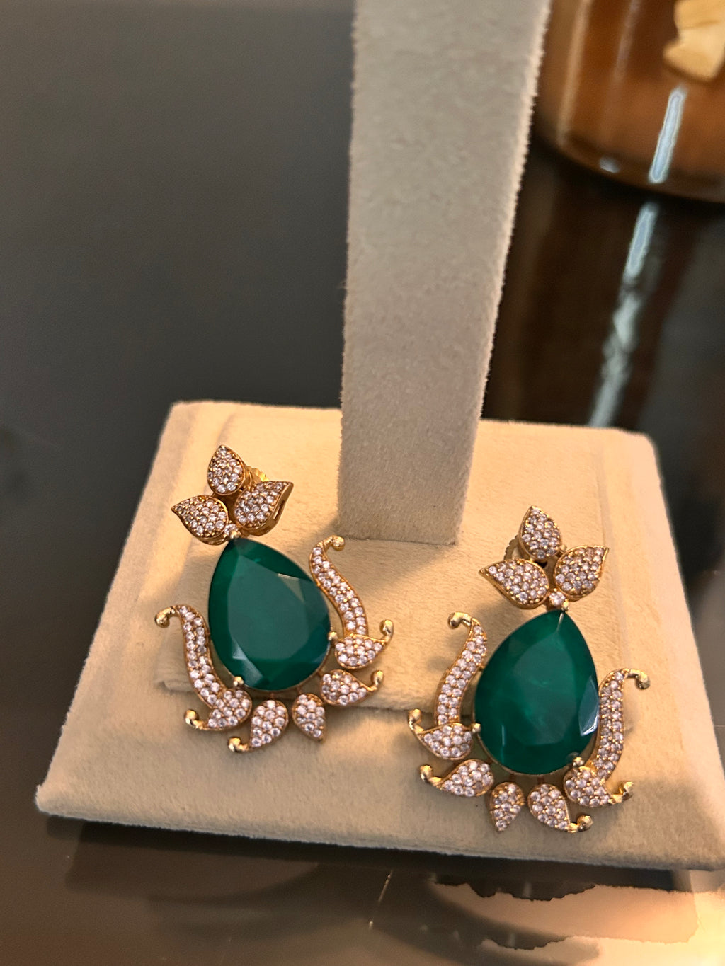 Gul earrings with green teardrop stones and clear-stone leaf details, shown as a pair at the base of a cream jewellery stand.