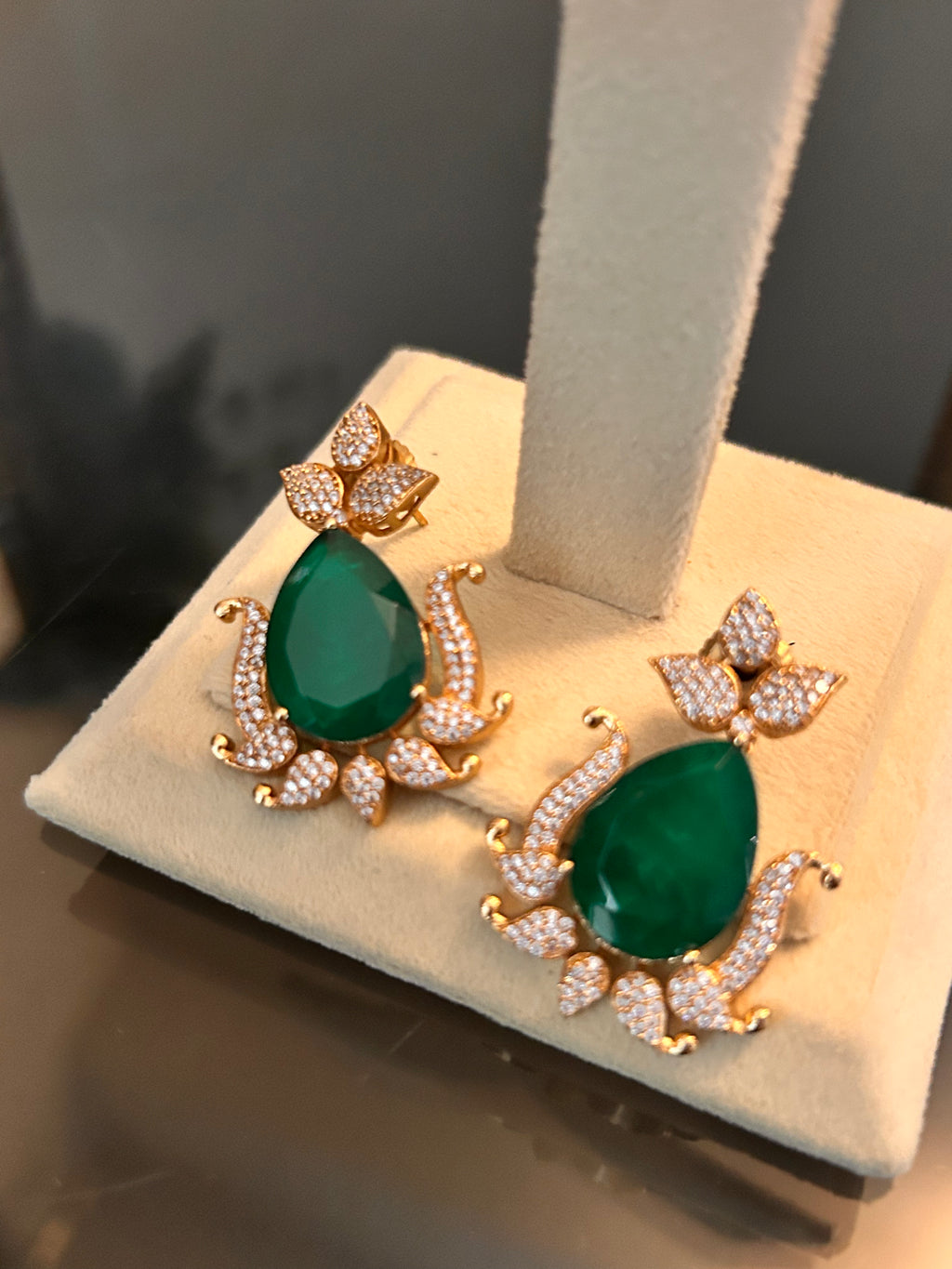 Angled close-up of Gul earrings resting on a cream stand base, highlighting the green stones and clear-stone leaf and scroll details.
