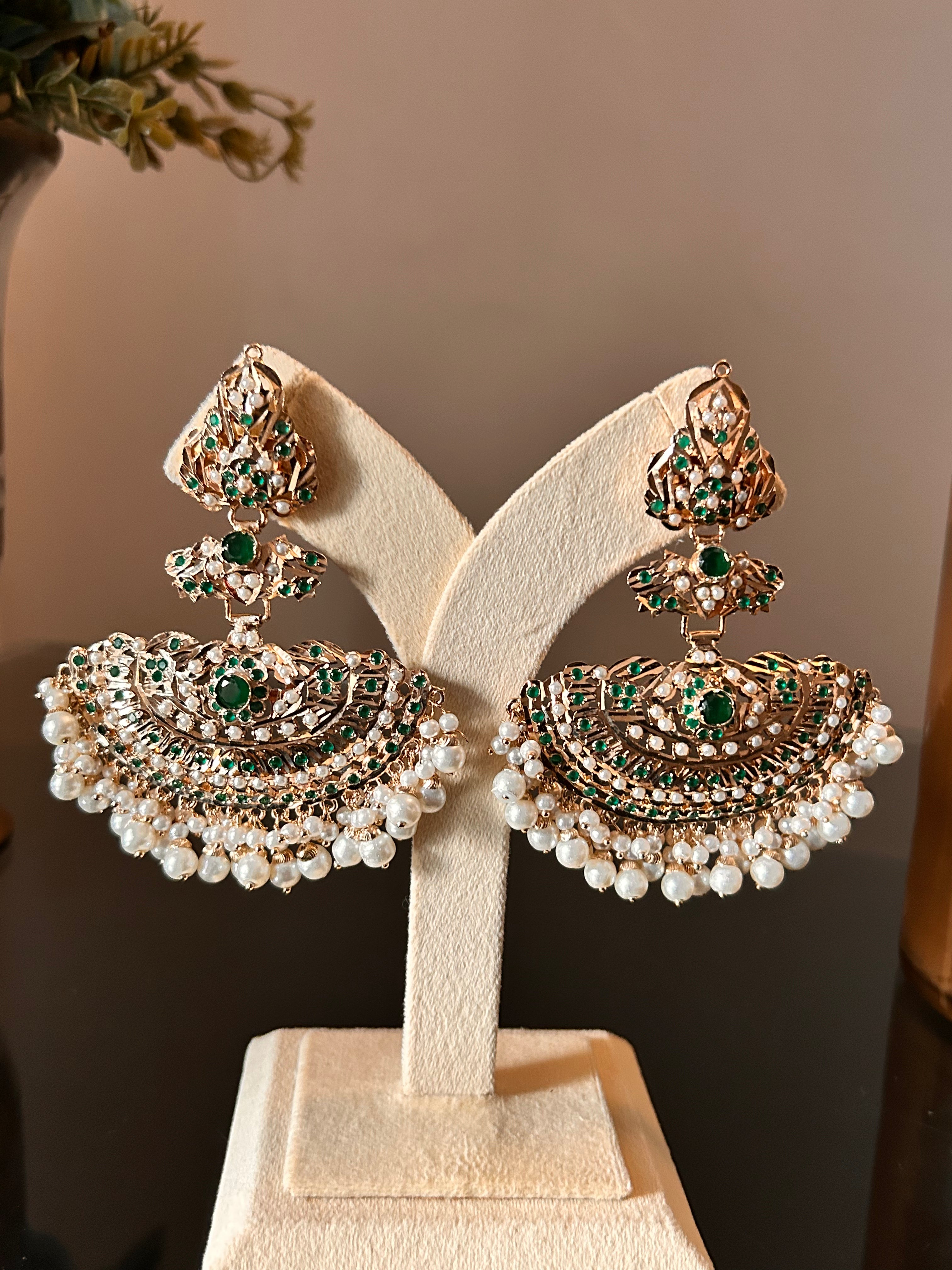 Khwahish earring pair hanging on a cream display against a dark background, with green and pink stones and pearl-like fringes.