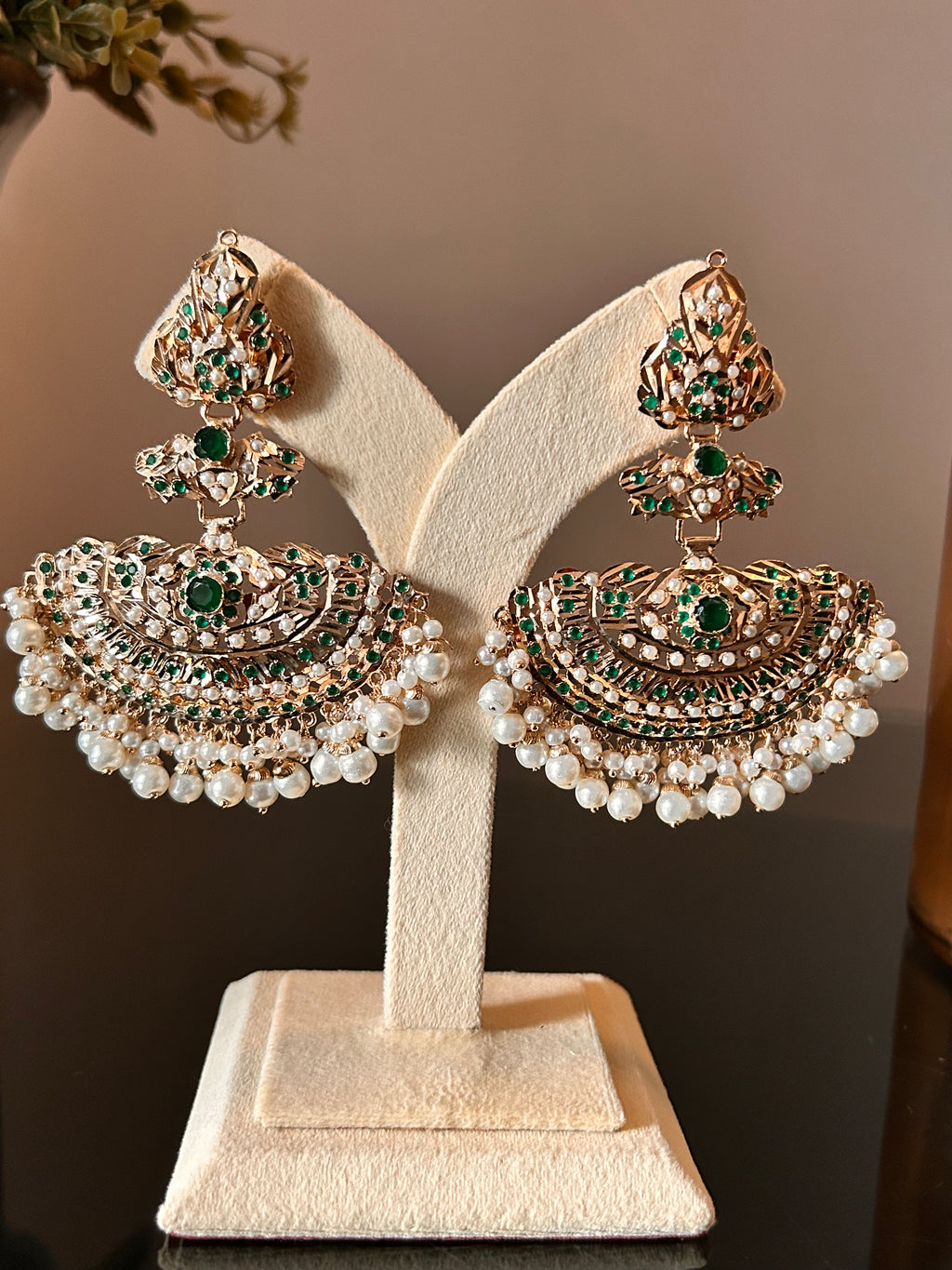 Close front view of Khwahish earrings hanging from a cream stand, with green and pink stones in ornate gold-tone settings.