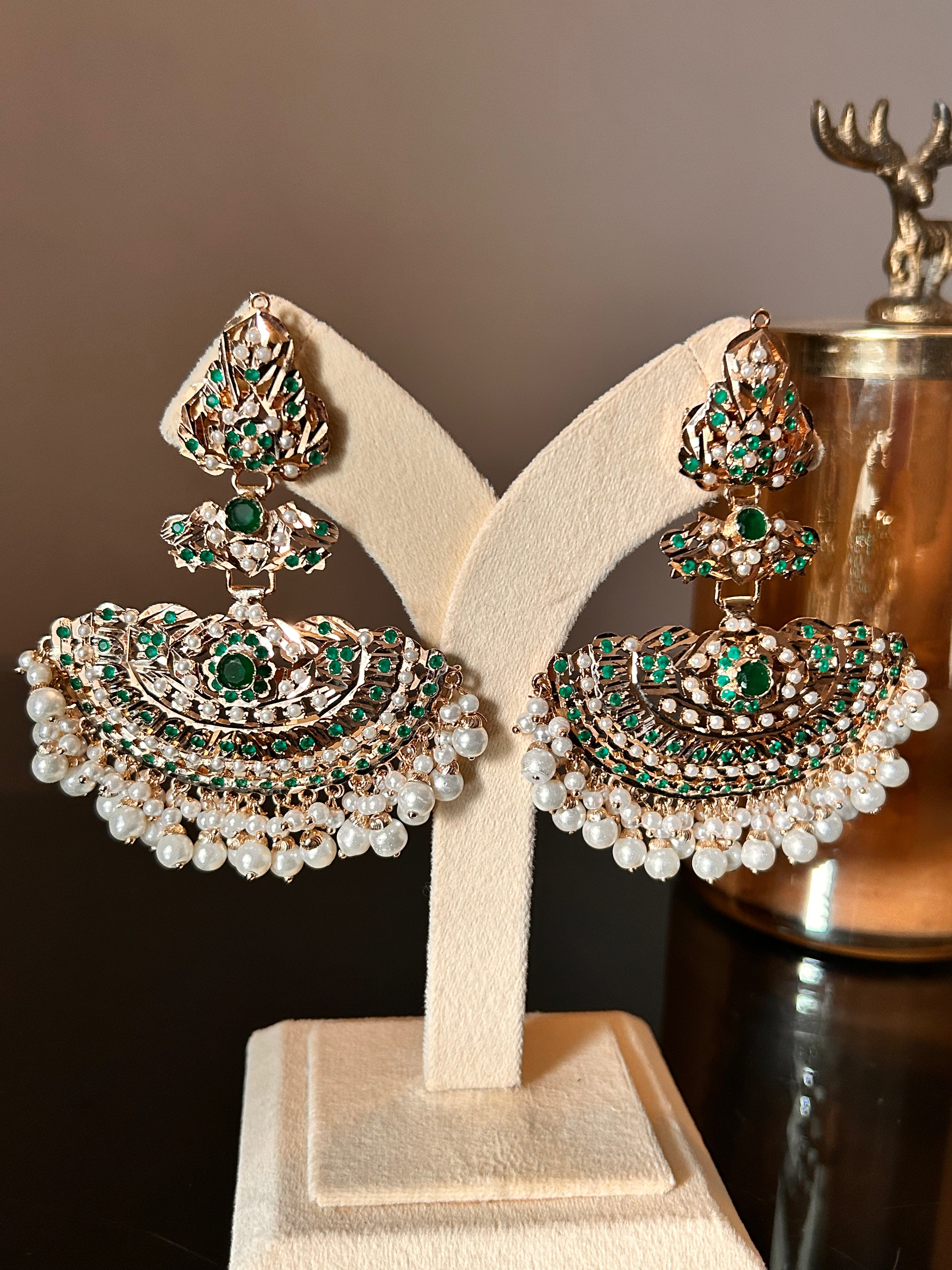 Angled view of Khwahish statement earrings on a cream stand, showing the layered green and pink stone motifs and pearl-like fringes.