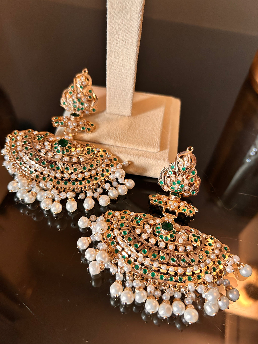 Close view of Khwahish earrings laid on a cream stand base, highlighting green and pink stones and the pearl-like fringe.