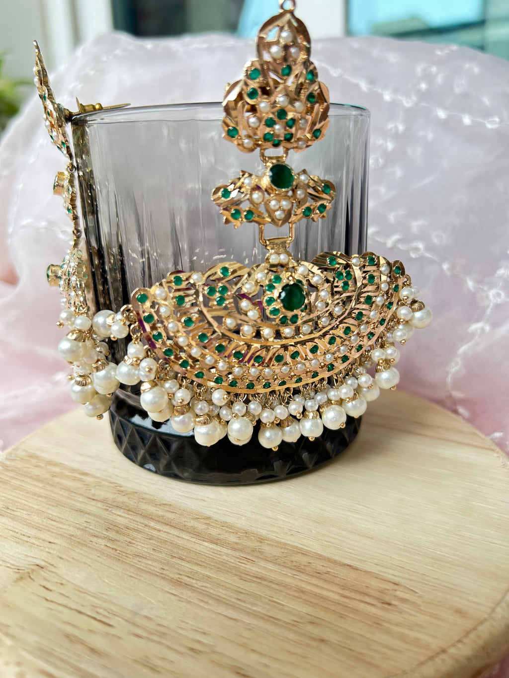 Angled close-up of a Khwahish earring on a glass, showing green and pink stonework, gold-tone details and pearl-like drops.