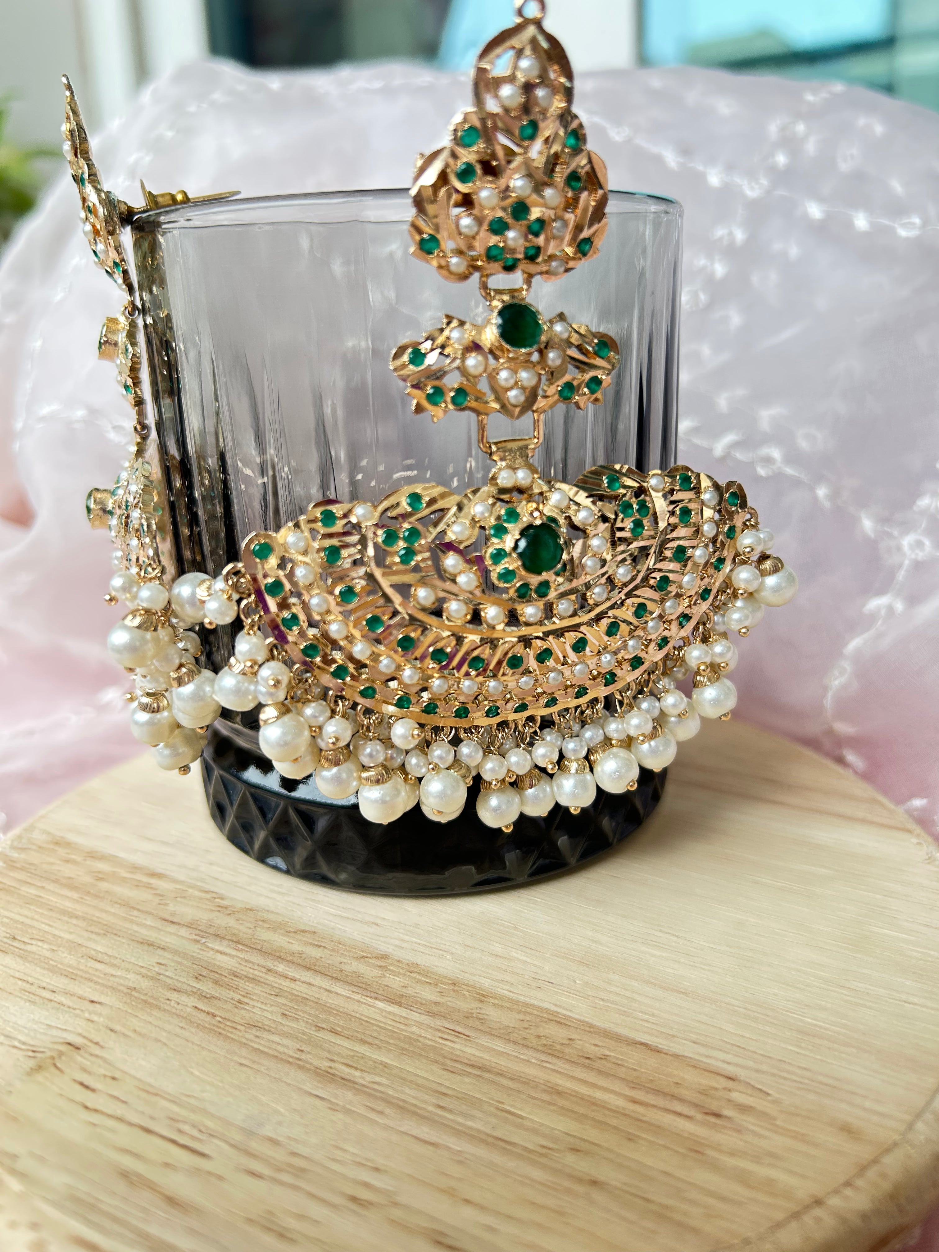 Angled close-up of a Khwahish earring on a glass, showing green and pink stonework, gold-tone details and pearl-like drops.