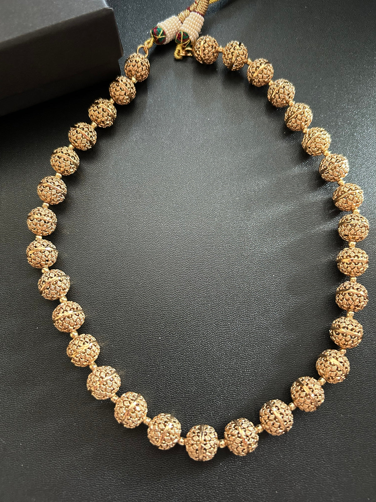Maira brass carved-bead necklace arranged on a dark surface beside a KINAHH box, showing the textured round beads.