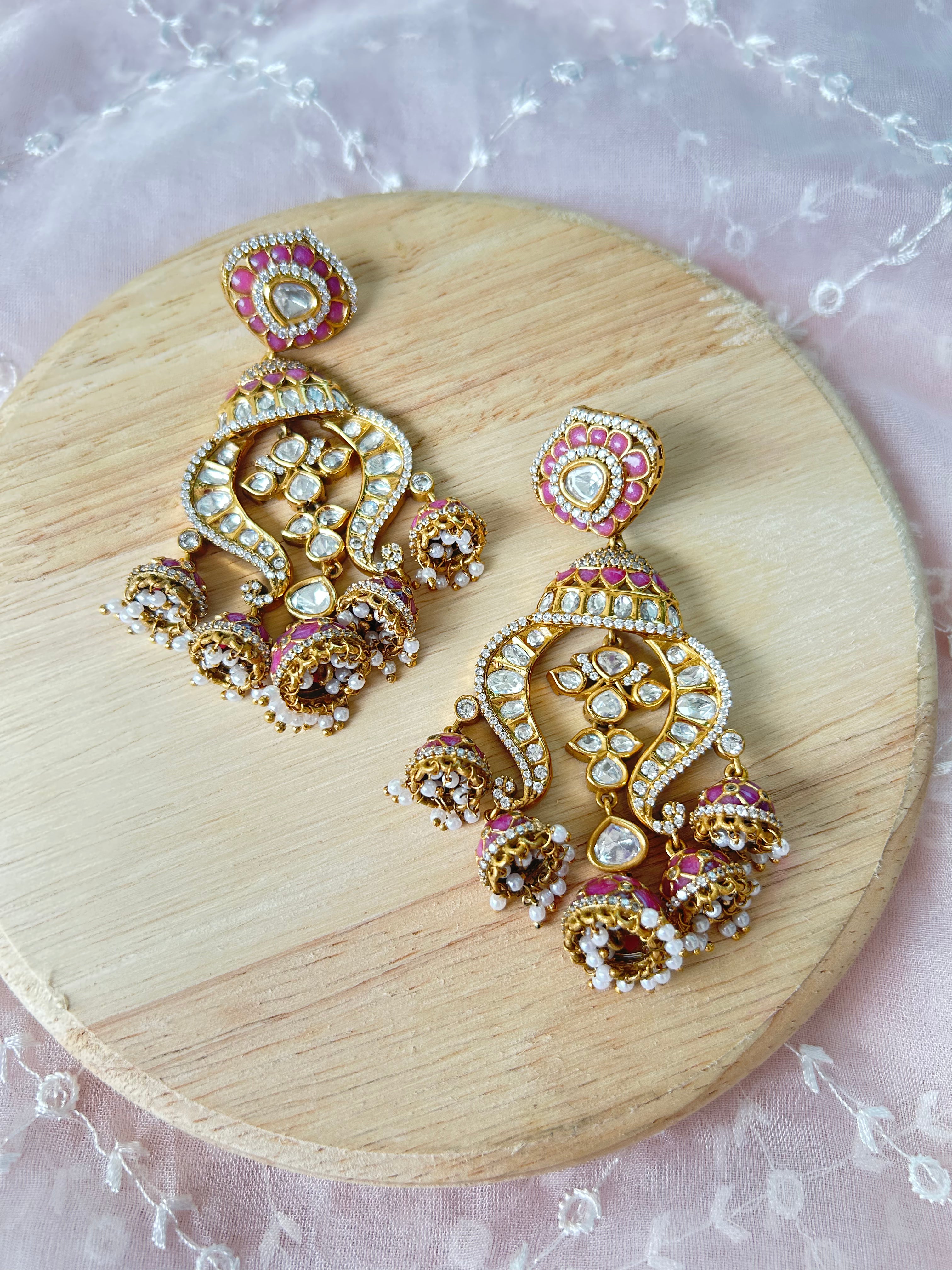 Alternate front view of Noor pink-enamel chandelier earrings, showing clear polki stones and dangling decorative details on a wooden display.