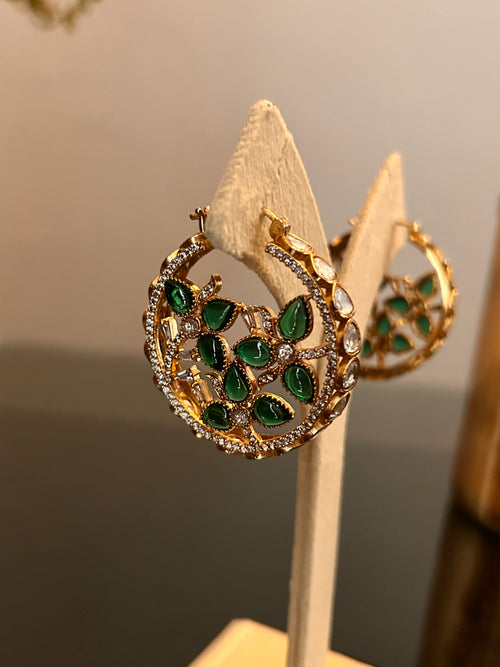 Angled close view of Chaand circular hoop earrings on a cream stand, with green teardrop stones and clear stone details.