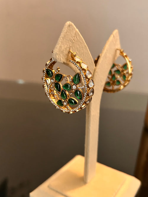 Chaand Earrings by KINAHH, shown as a pair on a cream stand with green teardrop stones and clear stone detailing.