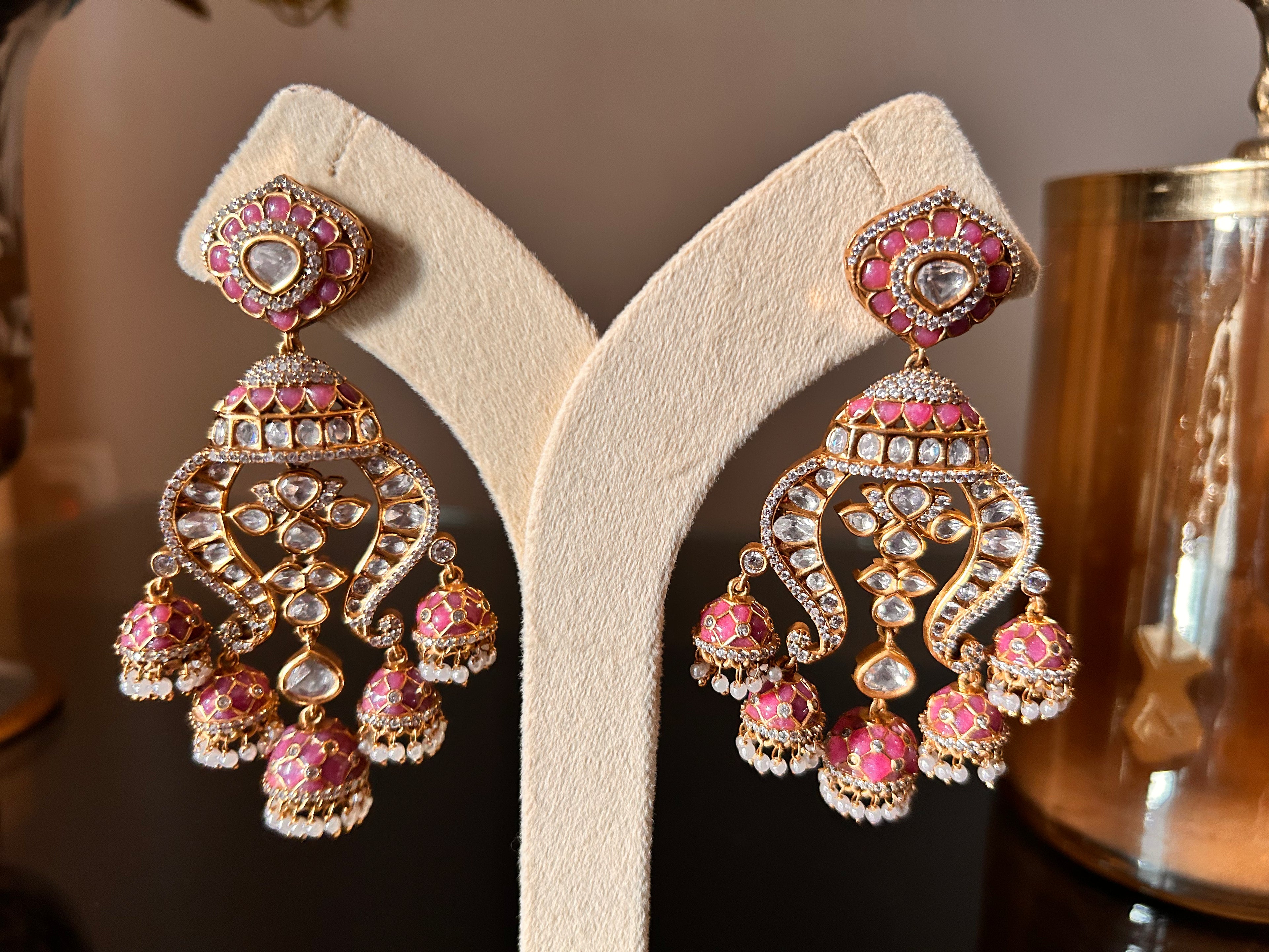 Close front view of Noor Earrings with pink enamel, clear stones and pearl-like drops, displayed as a pair on a cream stand.