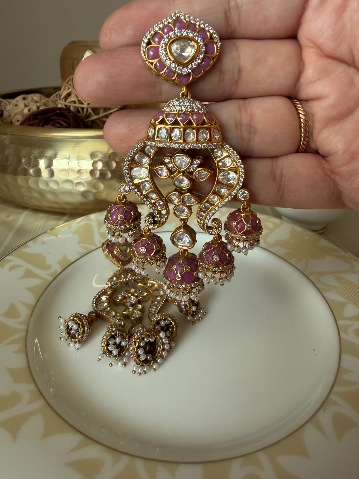 Noor earring held in hand, showing pink enamel, clear stones and pearl-like drops, with the matching earring behind.
